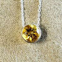 A round faceted citrine pendant bezel-set in sterling silver, displayed on an adjustable sterling silver chain against a beige background.