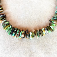 A close-up of a chunky beaded necklace made of polished natural stone chips in earthy green, brown, black, and white marbled patterns, displayed on a textured beige fabric background.